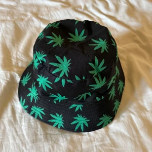 Svart bucket hat med grönt bladmönster - Cool svart bucket hat med gröna bladmotiv över hela hatten. Hatten har en klassisk form med nedvikt brätte och är tillverkad i ett mjukt tygmaterial. Perfekt accessoar för en avslappnad stil.