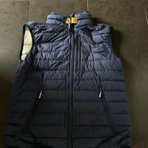 Blå dunväst från Parajumpers - Snygg blå dunväst från Parajumpers med hög krage och dragkedja framtill. Västen har tre fickor med dragkedja och en gul detalj vid halsen. Lätt och smidig modell, perfekt för lager-på-lager.