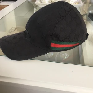 Svart keps från Gucci med logomönster - Säljer en svart keps från Gucci med diskret GG-logomönster och klassiska gröna och röda ränder på sidan. Kepsen har böjd skärm och justerbar rem baktill. Perfekt för dig som gillar exklusiva accessoarer.