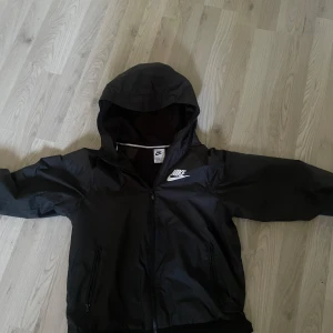 Svart vindjacka från Nike - Säljer en svart vindjacka från Nike med huva och dragkedja framtill. Jackan har två fickor med dragkedja och en vit Nike-logga på bröstet. Perfekt för blåsiga dagar och enkel att matcha med det mesta.