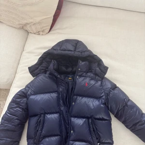 Mörkblå dunjacka från Polo Ralph Lauren - Säljer en mörkblå pufferjacka från Polo Ralph Lauren med huva och dragkedja. Jackan har det klassiska röda Polo-logot broderat på bröstet och är vadderad för extra värme. Perfekt för kyliga dagar. Väldigt fin och knappast använd skriv i dm om fler foton har själva lapparna kvar💗Den är från 12-14år och i polo storlek L