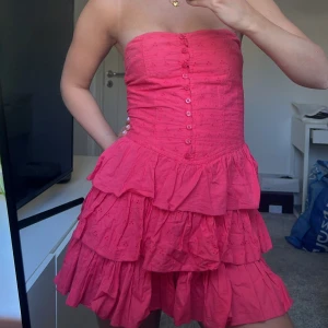 volangklänning - Säljer en söt axelbandslös rosa klänning med knappar framtill och flera volanglager. Klänningen har broderade detaljer och smock i ryggen för en bekväm passform. Perfekt för sommarens alla tillfällen!