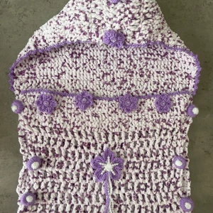 Handvirkad lila och vit babypåse med huva och blommor - Superfin handvirkad babypåse i lila och vitt med huva och dekorativa virkade blommor. Den har handvirkade knappar längs sidorna och en stor blomma framtill. Går att beställa andra färger tar ca 1/2 dagar