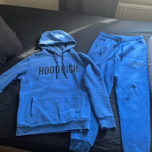 Blå dress från Hoodrich - Säljer en blåa dress från Hoodrich med svart logga på bröstet. Dressen har huva med dragsko, magficka och ribbade muddar. Materialet är mjukt och känns som bomull eller polyester. Perfekt för en avslappnad stil.