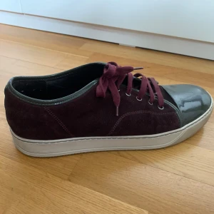 Vinröda sneakers från Lanvin med grå tå - Säljer ett par snygga vinröda sneakers från Lanvin med grå lackad tå och matchande vinröda skosnören. Skorna är riktigt fräscha i fint skick. Tveka inte att höra av er vid intresse eller vid fler bilder av skon. Dustbag och skolåda medkommer inte. Hör av er vid intresse