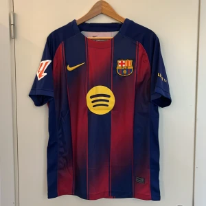 FC Barcelona hemmatröja Lamine Yamal 25/26 - Säljer en officiell FC Barcelona hemmatröja med Lamine Yamal och nummer 19 på ryggen. Tröjan är blå och röd med gula detaljer, klubbmärke på bröstet och sponsorloggor. Kortärmad modell i lätt och ventilerande material.