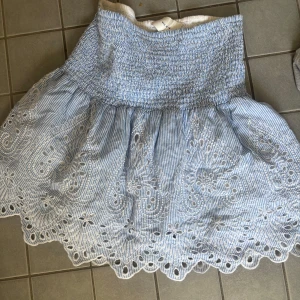 Blåvit kjol med broderade detaljer - Supersöt blåvit kjol med tunna ränder och broderade blommönster. Kjolen har en smockad midja och vågig kant nertill. Perfekt för varma dagar och ger en fräsch sommarkänsla.