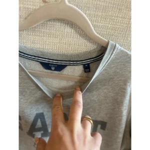 Grå sweatshirt från GANT - Klassisk grå sweatshirt från GANT med tryckt logga och text 'Est. 1949' på bröstet. Tröjan har rund halsringning och långa ärmar. Perfekt för en avslappnad och stilren look.