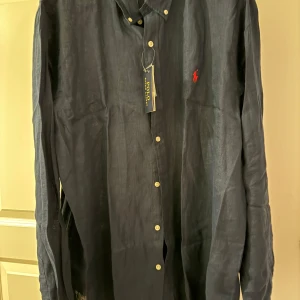 Mörkblå linneskjorta från Polo Ralph Lauren - Snygg mörkblå skjorta i linne från Polo Ralph Lauren. Slim fit-modell med klassisk krage, vita knappar och den ikoniska röda loggan broderad på bröstet. Perfekt för dig som gillar stilrena och tidlösa plagg.