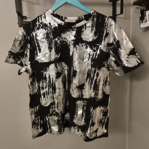 Svart t-shirt med silvrigt mönster - Cool svart t-shirt med unikt silvrigt abstrakt mönster. T-shirten har rund hals och korta ärmar, perfekt för dig som vill sticka ut med en modern och edgy stil.