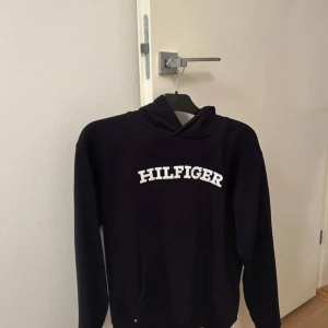 Mörkblå hoodie från Tommy Hilfiger - Säljer en blå hoodie från Tommy Hilfiger med vit broderad logga på bröstet. Klassisk huva och känguruficka framtill. Perfekt för en avslappnad och stilren look.