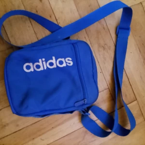 Blå axelväska från Adidas - Säljer en blå axelväska från Adidas med vit logga framtill. Väskan har justerbar axelrem och ett fack med dragkedja. Perfekt för att bära med sig det viktigaste till vardags eller på språng.