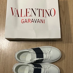 Valentino Garavani vita sneakers med mörkblått band - Säljer ett par snygga vita sneakers från Valentino Garavani med ett brett svart band över vristen och vita nitar på sidorna. Skorna har rund tå, platt sula och klassisk snörning. Perfekta för dig som gillar stilrena och exklusiva sneakers.