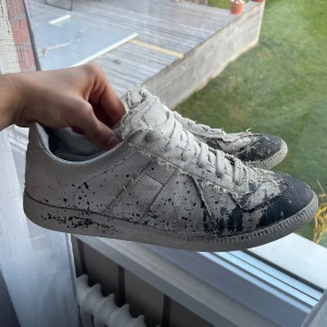 Maison Margiela Gats - Unika sneakers från Maison Margiela i en vit nyans med svart färgstänk. Skorna har en rund tå och snörning framtill. Perfekta för dig som vill ha en konstnärlig touch på din stil. Box och dustbag tillkommer. Finns kvitto. Inte bästa skicket därav priset. Finns defekter i sulan och i skorna men det är inget man märker vid användning.