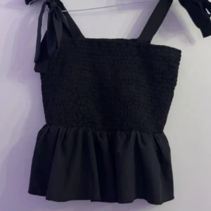 Svart smockad linnetopp med knytband från SHEIN - Supersöt svart linnetopp från SHEIN med smockad överdel och volang nertill. Axelbanden knyts i rosetter på axlarna för en extra gullig touch. Perfekt till sommaren och lätt att matcha med jeans eller kjol.