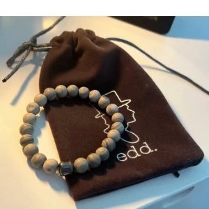 Armband med gråmelerade träpärlor från Edd - Snyggt armband med runda gråmelerade träpärlor och en metallfärgad detalj. Kommer i en brun tygpåse med Edd-logga. Perfekt accessoar för en stilren look.