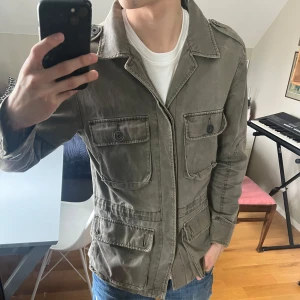 Grön Fieldjacka - Säljer en grön jacka med fyra stora fickor framtill och klassisk krage. Jackan har knappdetaljer och dragkedja, tillverkad i bomull. Perfekt för dig som gillar en avslappnad och stilren look.