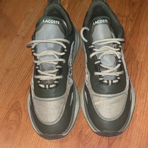 Gröna och beige sneakers från Lacoste - Säljer ett par snygga sneakers från Lacoste i grönt och beige med tydlig logga på sidan och hälen. Skorna har snörning, rund tå och en platt sula med mönster undertill. Perfekta för en sportig och avslappnad stil. Använda 5-10 gånger max. Köpte för 1500. Jag rengör dem innan jag fraktar så att dem ser nya ut! 