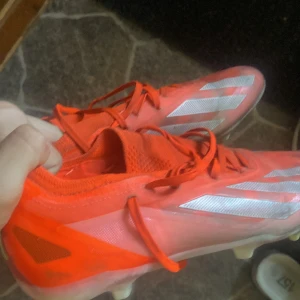 Röda fotbollsskor från Adidas - Säljer ett par röda Adidas fotbollsskor med vita detaljer och snörning. Skorna har en låg profil och är designade för bra grepp på planen. Perfekta för dig som vill sticka ut på matchen! Fotbolllskornaa är rätt trasiga och hål på många ställen.