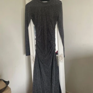 Glittrig långklänning från Kappahl - Säljer en snygg långärmad glittrig långklänning från Kappahl i svart med silverton. Klänningen har rund halsringning och rynkade detaljer längs sidan för en smickrande passform. Perfekt för dig som vill sticka ut.