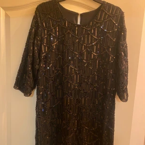 Svart paljettklänning med mönster - Säljer en svart klänning täckt av paljetter och glittriga detaljer i geometriskt mönster. Klänningen har rund halsringning och trekvartsärm, perfekt för att sticka ut på festen. Fodrad insida för extra komfort. 