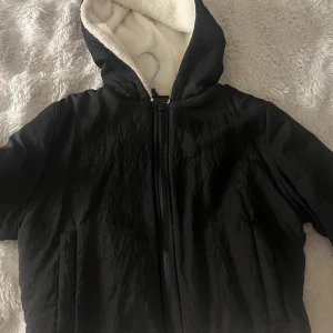 Svart pufferjacka med fuskpälsfoder - Mysig svart pufferjacka med huva och vitt fuskpälsfoder. Jackan har dragkedja framtill och är perfekt för kyliga dagar. Stilen är enkel och modern med lång ärm och croppad passform.