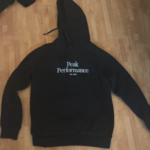 Svart hoodie från Peak Performance - Säljer en svart hoodie från Peak Performance med vit logga på bröstet. Klassisk modell med huva och magficka, perfekt för en avslappnad stil. Tjockt och mjukt material som håller dig varm under kyliga dagar.Den är i storlek M men lite liten i storleken.