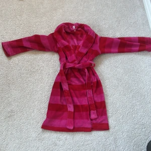 Röd och rosa randig morgonrock med bälte - Mysig morgonrock i röd och rosa med breda ränder och knytskärp i midjan. Långärmad modell med stor krage och mjukt material, perfekt för att hålla sig varm hemma.