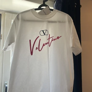 Vit t-shirt från Valentino - Vit t-shirt från Valentino med röd logotyp och tryck framtill. Klassisk rund halsringning och korta ärmar. Perfekt för en stilren outfit till sommaren. Oanvänd!