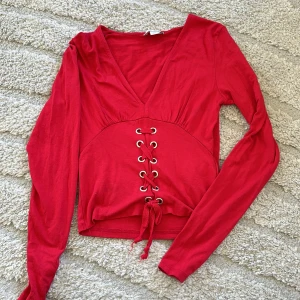 Röd långärmad topp med snörning - Trendig röd långärmad topp med djup v-ringning och snygg snörningsdetalj framtill. Croppad modell som passar perfekt till både jeans och kjol. Mjuk och skön bomullskänsla.