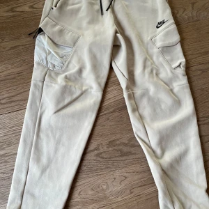 Beige mjukisbyxor från Nike - Säljer ett par beige mjukisbyxor från Nike med svarta dragsnören och flera fickor, inklusive en med lock. Byxorna har en avslappnad passform och är perfekta för en chill stil. Nike-logga broderad på ena benet. Lite noppiga på baksidan, se bild.
