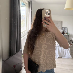 Beige mönstrad blus med swirl-detaljer - Säljer en beige blus med swirl-mönster i ett strukturmaterial. Blusen har korta ärmar och en lös passform, perfekt till jeans eller kjol. Materialet känns mjukt och luftigt, och toppen har en rund halsringning.