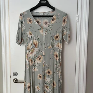 Blommig långklänning från H&M - Ljusgrön långklänning från H&M med blommigt mönster i beige, brunt och vitt. Klänningen har v-ringning och korta ärmar, samt en mjuk och flowy passform. Bra skick eftersom den använts fåtal gånger!