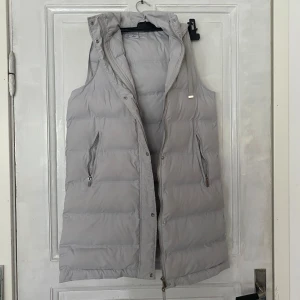 Ljusgrå dunväst från Stories - Ljusgrå dunväst från Stories med huva och dragkedja framtill. Västen har två fickor med dragkedja och quiltad design. Perfekt för lager-på-lager och enkel att styla till olika outfits.