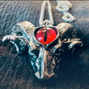 Halsband med draköga och horn – satanic, metal, goth/alternative - Tungt halsband i rostfritt stål med rå amulett: bockhuvud med böjda horn och intensivt rött draköga i glas. Kedjan är 60 cm lång och stängs med karbinhake. Amuletten mäter ca 4,5 × 4,5 cm. Helt nytt och oanvänt – passar alla kön. Ingen prutning, men bundlerabatt ges. Rensar smyckesskrin och garderob – kika gärna på mina andra annonser också!