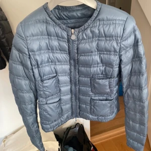 Ljusblå pufferjacka från Moncler - Säljer en ljusblå pufferjacka från Moncler med rund halsringning och dragkedja framtill. Jackan har två stora fickor framtill och är lätt vadderad, perfekt för övergångssäsonger. Klassisk Moncler-logga på ärmen.