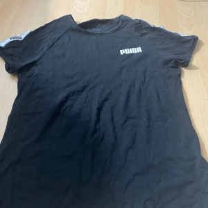 Svart t-shirt från Puma - Säljer en svart t-shirt från Puma med logga på bröstet och vita detaljer på ärmarna. Klassisk passform och rund halsringning. Perfekt för en sportig stil eller till vardags. Materialet är mjuk bomull.