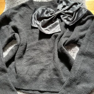 Svart stickad tröja med stor rosett från Kappahl - Snygg svart stickad tröja från Kappahl med en stor, glansig rosett på axeln. Tröjan är även lite nopprig. Tröjan har lång ärm och rund halsringning. Så elegant, perfekt till en sommar middag