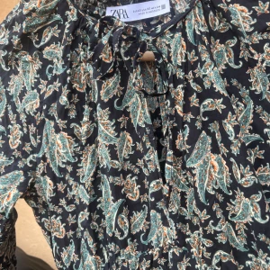 Svart mönstrad klänning från Zara - Svart klänning från Zara med grönt och orange paisleymönster. Klänningen har långa ärmar, knytning vid halsen och lätt rynkade detaljer. 