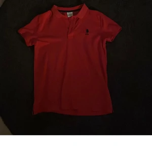 Röd pikétröja från U.S. Polo Assn. - Säljer en klassisk röd pikétröja från U.S. Polo Assn. med kort ärm och liten broderad logga på bröstet. Perfekt för en sportig och stilren look. Pikékrage och knappar framtill.