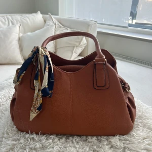 Brun handväska med scarfdetalj - Säljer en snygg brun handväska med matchande axelrem. Väskan är som helt ny och endast använd vid något enstaka tillfälle.Scarfen ingår inte 💕 