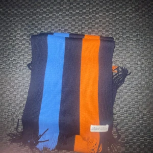 Randig halsduk med blå och orange ränder - Snygg halsduk med breda ränder i blått, svart och orange. Halsduken har fransar i ändarna och en liten tygpatch med märket Clint East. Perfekt accessoar för att sticka ut i mängden.