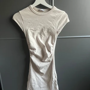Beige kortärmad klänning - Säljer en enkel och stilren beige kortärmad klänning med rund halsringning. Klänningen har en tight passform och är tillverkad i ett mjukt bomullsliknande material. Perfekt för en avslappnad och trendig look. Den är använd en gång och har draperingar på sidorna
