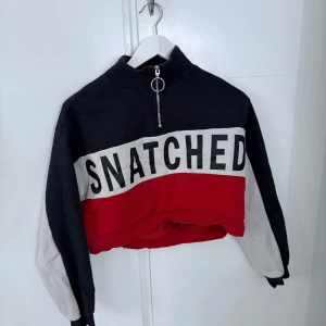 Svart, röd och vit croppad tröja från Divided - Säljer en croppad tröja från Divided med texten 'SNATCHED' framtill. Tröjan har halv dragkedja, höga muddar och färgblock i svart, rött och vitt. Perfekt för en trendig streetstyle-look.