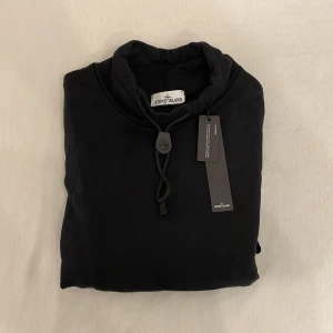 Svart sweatshirt från Stone Island (L) - Svart sweatshirt från Stone Island med hög krage och dragsko. Tröjan har långa ärmar, fickor med dragkedja och den klassiska logotypen på ärmen. Perfekt för en stilren och avslappnad look.