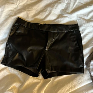 Svarta veganska skinn/läder shorts  - Snygga lågmidjade svarta shorts i veganskt skinn, de har normal passform, fickor i sidorna och stängs med dragkedja och hake. Perfekta för en trendig look och lätta att matcha med olika toppar.