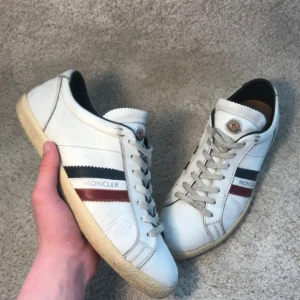 Vita sneakers från Moncler med röd och blå detalj - Säljer ett par vita Moncler sneakers med klassiska röd och blå ränder på sidan och Moncler-logga. Skorna har snörning, rund tå och platt sula. Tillverkade i skinn med bruna innersulor och diskret branding på hälen. Storlek 40 men passar mer som 41. Nypris 7000kr, mitt pris 1499kr. Bytte kan tillföras beroende på erbjudande 