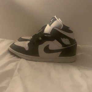 Svarta och vita Nike Air Jordans - Säljer ett par klassiska Nike Air Jordans i svart och vitt. Skorna har högt skaft, snörning och den ikoniska Air Jordan-loggan på sidan. Perfekta för dig som gillar streetwear och vill ha en tidlös sneaker i garderoben.