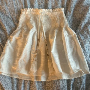 Söt kjol❤️ - Säljer en beige kjol med smock i midjan och dekorativa knytband med tofsar framtill. Kjolen har en lätt utsvängd form och fina detaljer i tyget som ger en bohemisk känsla.