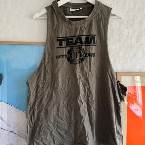 Olivgrön ärmlös linnetopp från Better Bodies - Säljer en olivgrön ärmlös linnetopp från Better Bodies med svart TEAM-tryck framtill. Perfekt för träning och gym, med lös passform och rund halsringning.